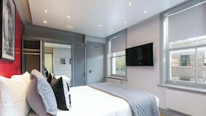 A modern bedroom with a large bed, a walk-in closet, a flat-screen TV, and a window with a city view. - The Bloomsbury - by Frankie (London)