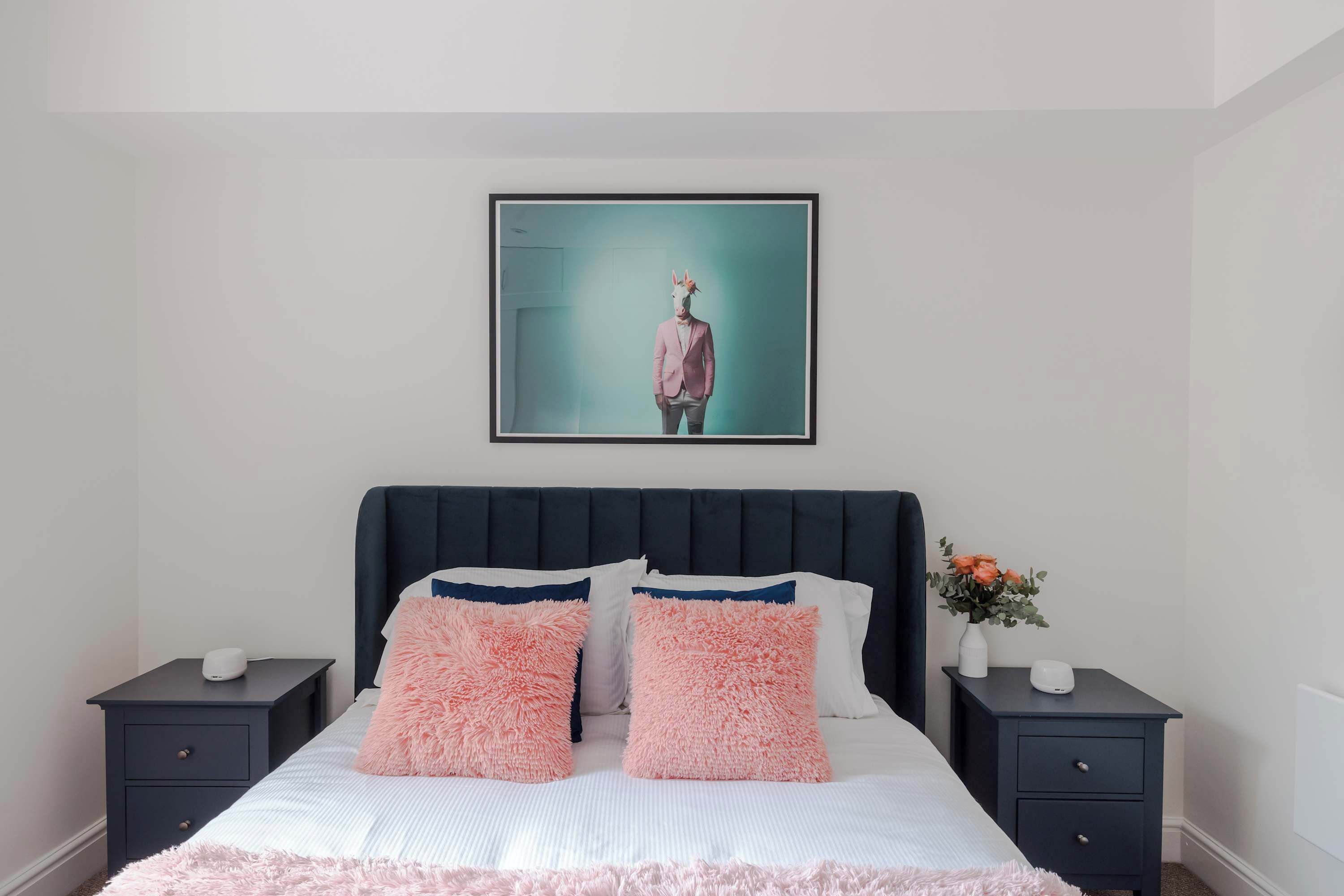Een slaapkamer met een bed, twee nachtkastjes, een ingelijst schilderij aan de muur en een vaas met bloemen.