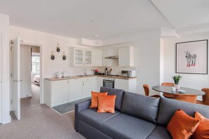 A modern kitchen with white cabinets, a dining area with a round table, and a gray sofa with orange cushions. - The Foley Residence - Fitzrovia - by Frankie (London)
