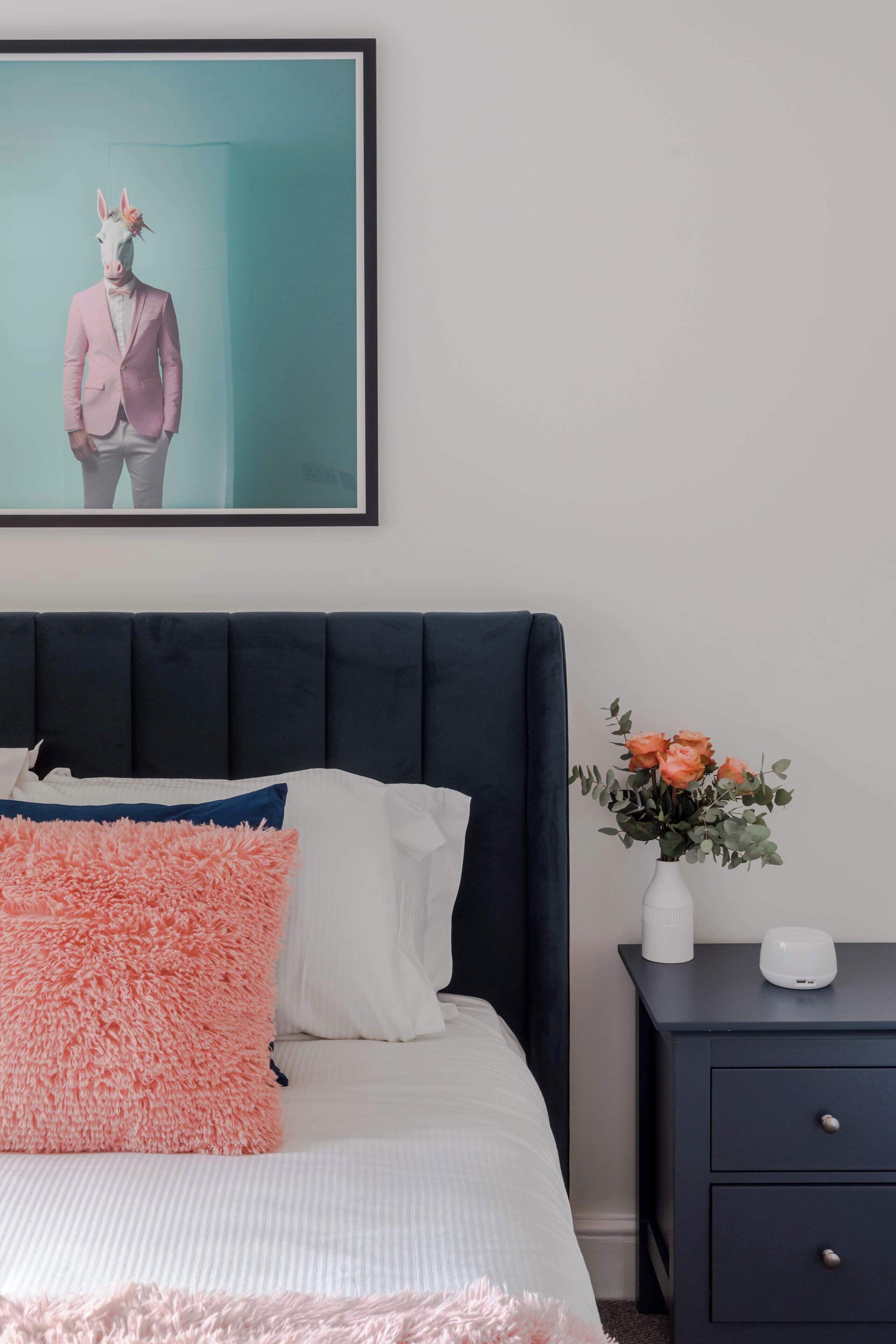 A bedroom with a bed, pillows, a nightstand, a vase with flowers, and a framed picture on the wall.