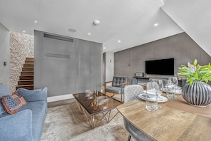 Een moderne woonkamer met een glazen salontafel, grijze banken, een eettafel met stoelen, een trap en een flatscreen televisie.