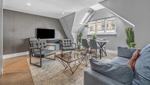 Een moderne woonkamer met een loft-plafond, een flatscreen-tv, grijze banken en een eethoek met een glazen salontafel.