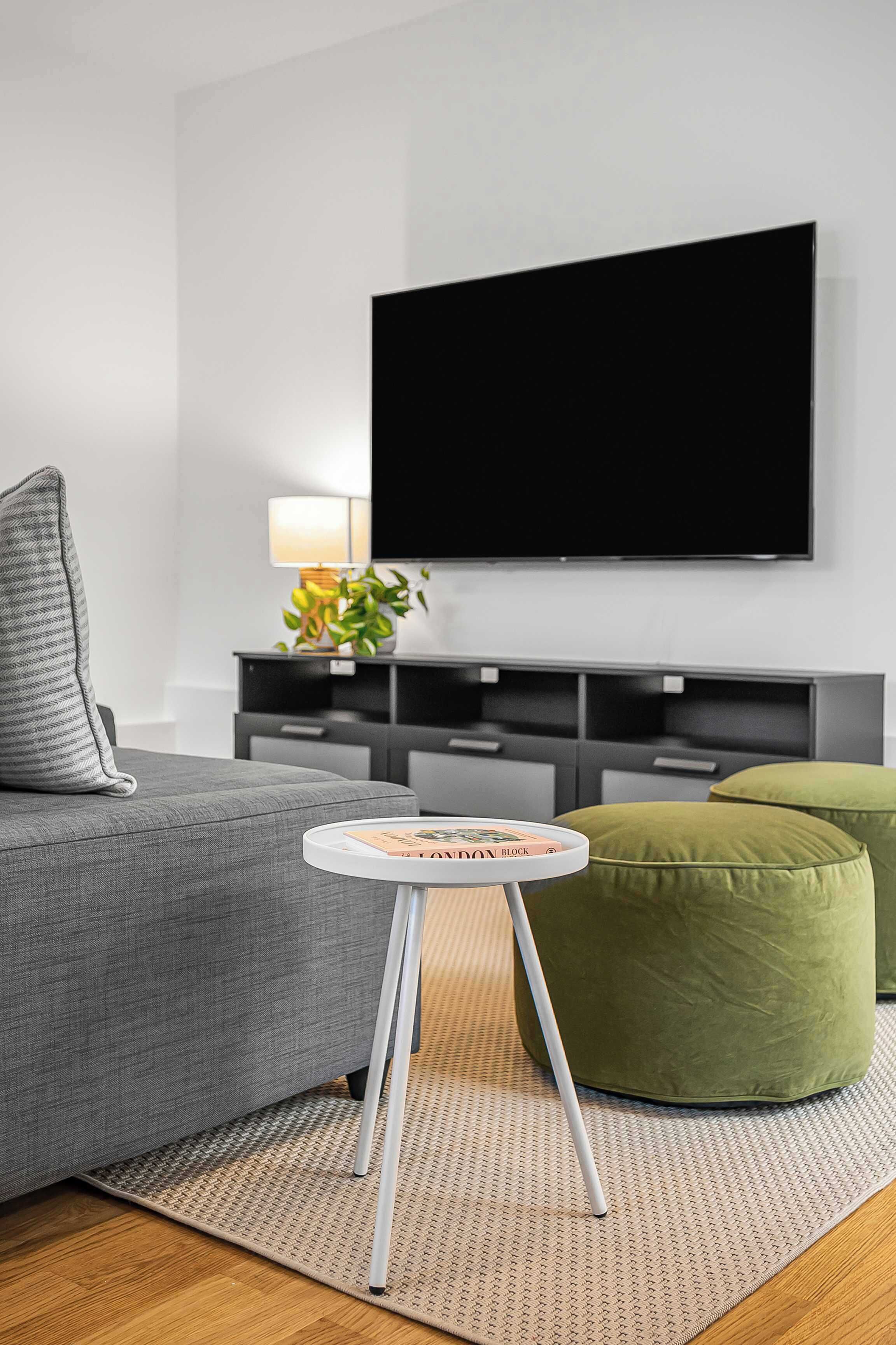 A modern living room with a grey sofa, a round white side table, a green ottoman, a flat-screen TV mounted on the wall, and a small plant on a side table.