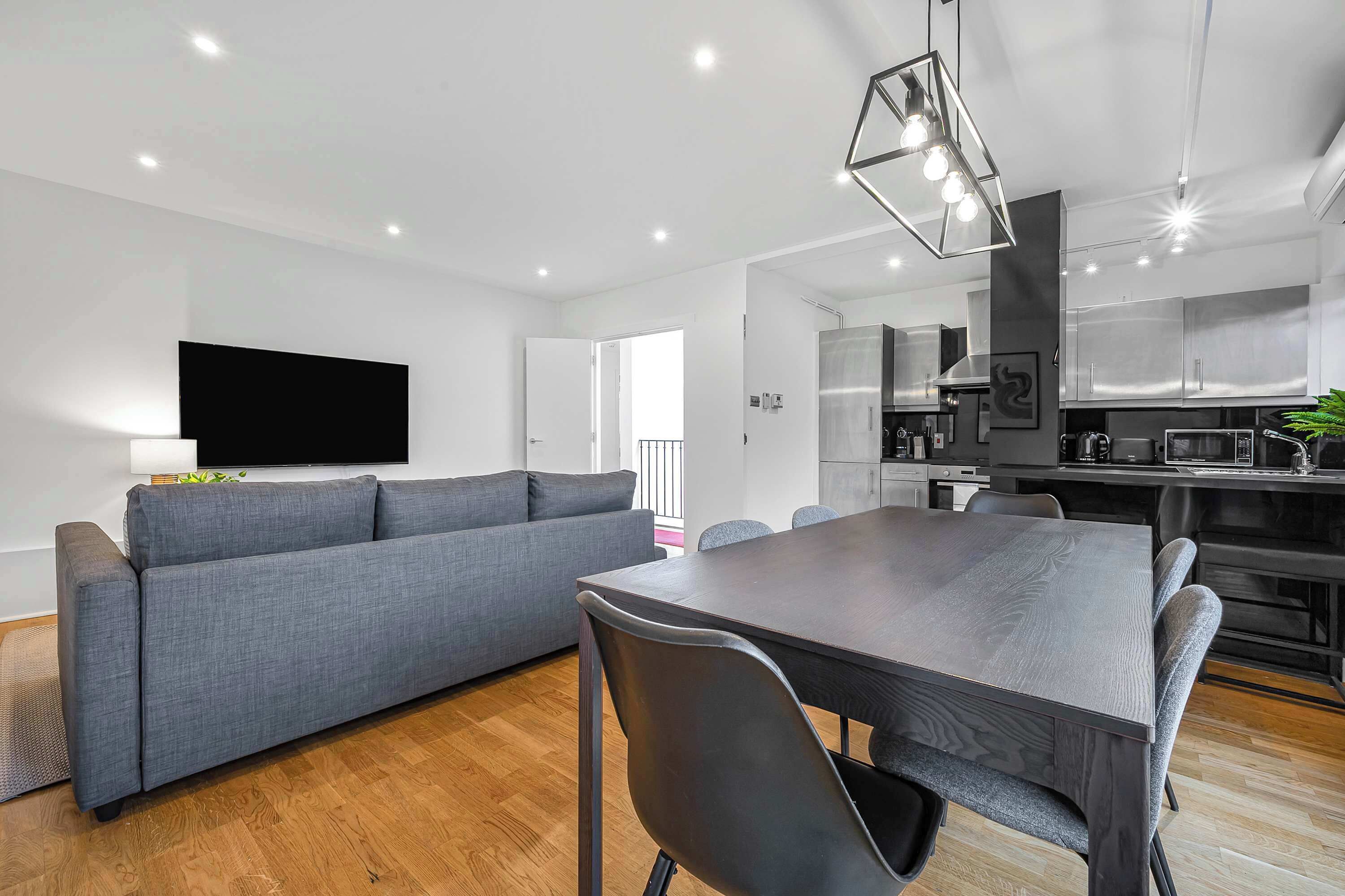 Ein modernes Wohnzimmer mit grauem Sofa, einem Esstisch mit Stühlen, einem wandmontierten Flachbildfernseher und einer Küchenzeile mit Edelstahlgeräten.