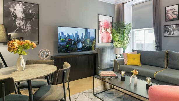 Una sala moderna con una televisión que muestra un paisaje urbano, una mesa de comedor con sillas, un sofá con cojines y una planta en maceta.
