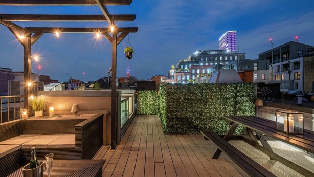 En takterrass med trägolv, en pergola med ljusslingor, en sittgrupp med kuddar, en krukväxt och utsikt över stadens byggnader.
