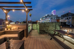 A rooftop terrace with wooden flooring, a pergola with string lights, a seating area with cushions, a potted plant, and a view of city buildings. - The Influencer Suite - Soho - by Frankie Says (London)