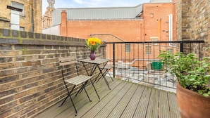 Ein Balkon mit Holzboden, Metallgeländer, einem Tisch mit Stühlen, einem Blumentopf und einer Backsteinmauer.