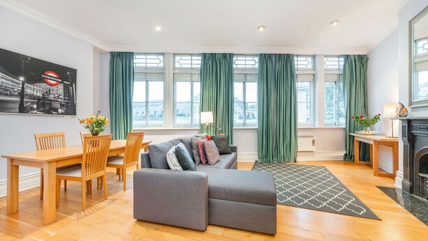 Ein modernes Wohnzimmer mit grauem Sofa, Esstisch aus Holz und Stühlen. Große Fenster mit grünen Vorhängen, ein Kamin und ein Bild an der Wand.