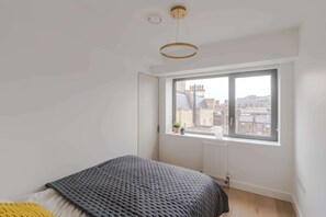 A bedroom with a large window, a bed, a radiator, and a hanging light fixture. - The Portland Penthouse - Marylebone - by Frankie (London)