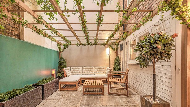 A cozy outdoor seating area with a sofa, two wooden tables, and a planter with a tree. - Fitzroy Garden - Fitzrovia - by Frankie (London)