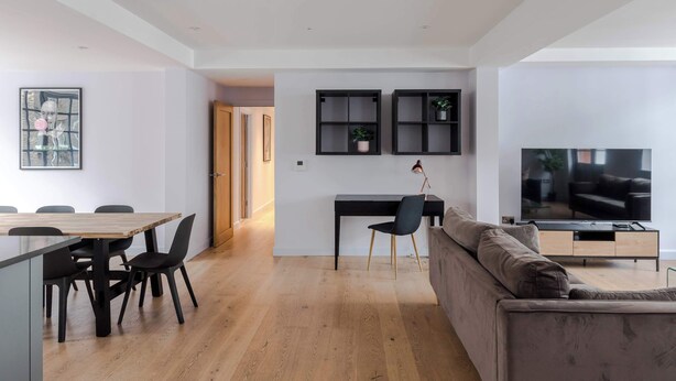 A modern living room with a wooden dining table, black chairs, a grey sofa, a TV, and built-in shelves with plants. - The Eyre Suite Two - Farringdon - by Frankie (London)
