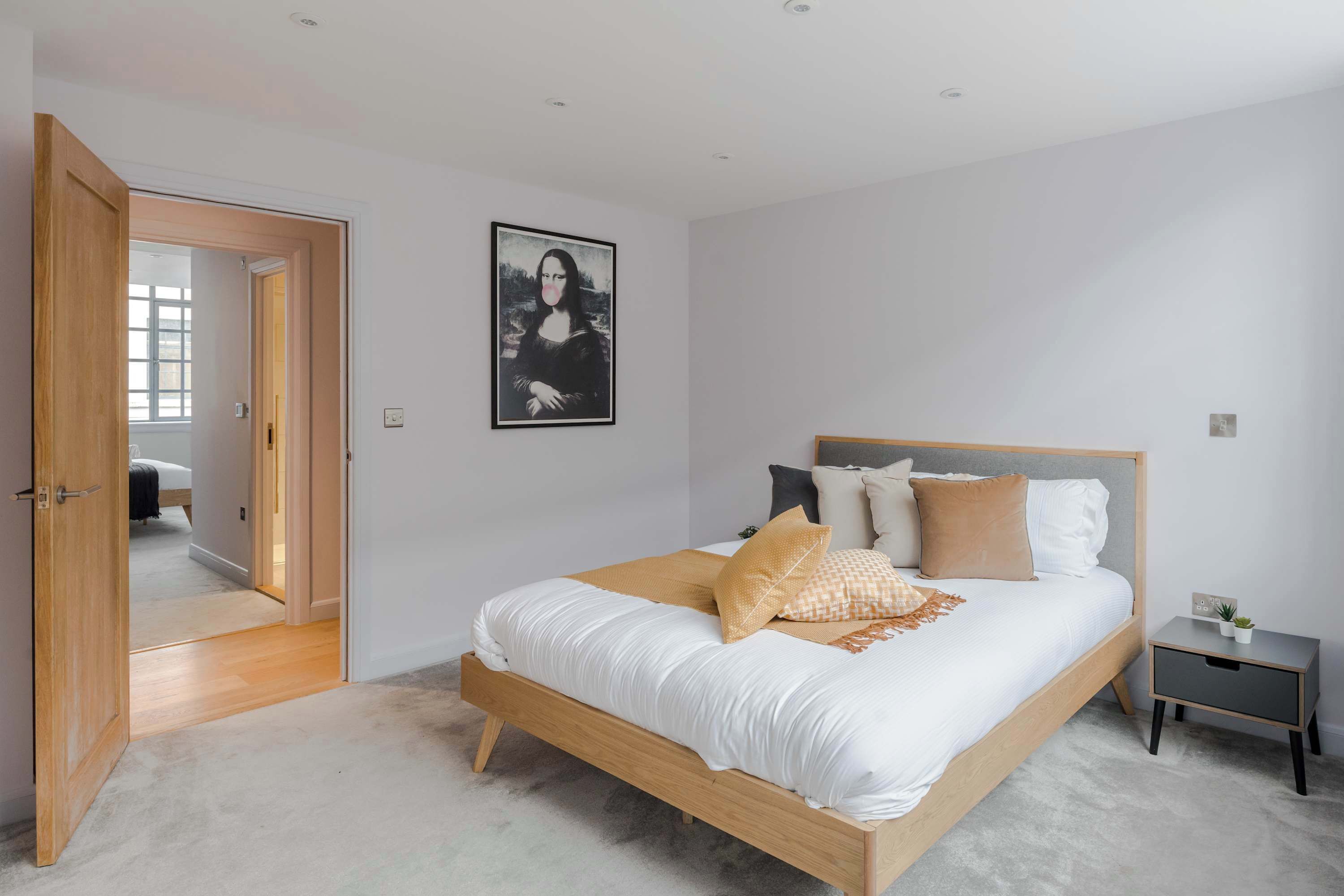 A bedroom with a wooden bed, a bedside table, a nightstand, a framed artwork on the wall, and a door leading to another room.