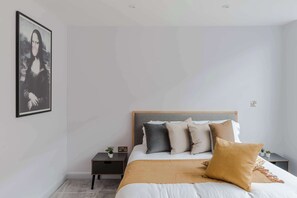 A bedroom with a bed, pillows, a nightstand, a wall-mounted picture, and a small plant. - The Eyre Suite Two - Farringdon - by Frankie (London)