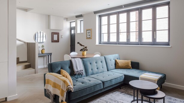 A modern living room with a sectional sofa, a small round table, and a large window with blinds. - The Tranquil Den - Regent's Park - by Frankie Says (London)