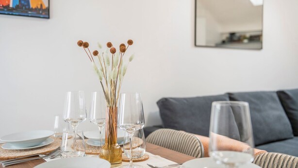 A dining table set with clear glassware and plates, a vase with dried flowers, and a grey sofa in the background. - The Cutesy - Fitzrovia - by Frankie Says (London)