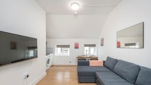 A modern living room with a grey sectional sofa, a flat-screen TV mounted on the wall, a washing machine, and a dining area with a table and chairs. - The Cutesy - Fitzrovia - by Frankie Says (London)
