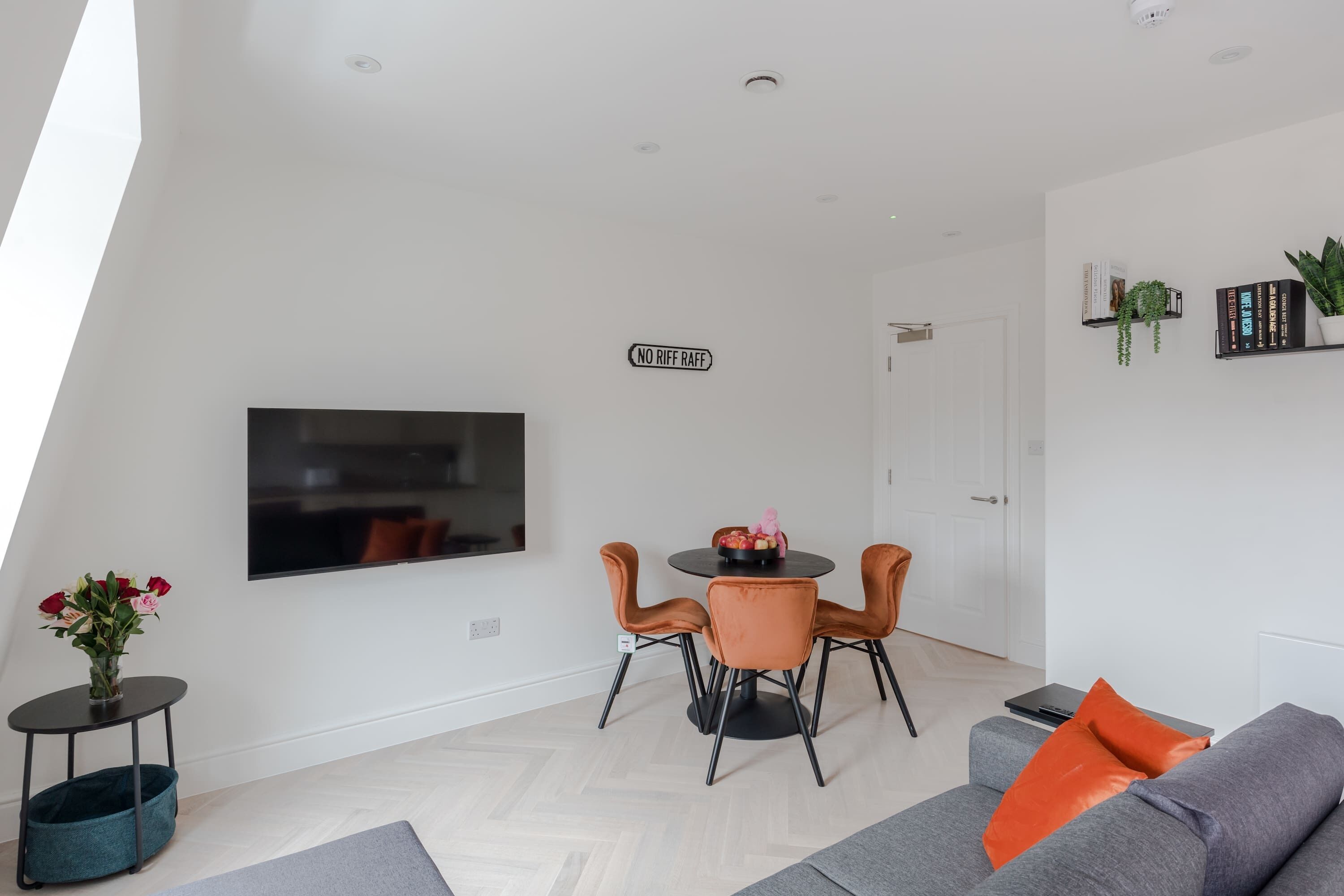 A modern living room with a flat-screen TV, a dining table with chairs, a grey sofa, and a small side table with a vase of flowers.