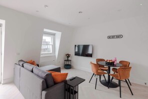 Uma sala de estar moderna com sofá, mesa de jantar com cadeiras, TV fixada na parede e uma pequena mesa lateral com um vaso.