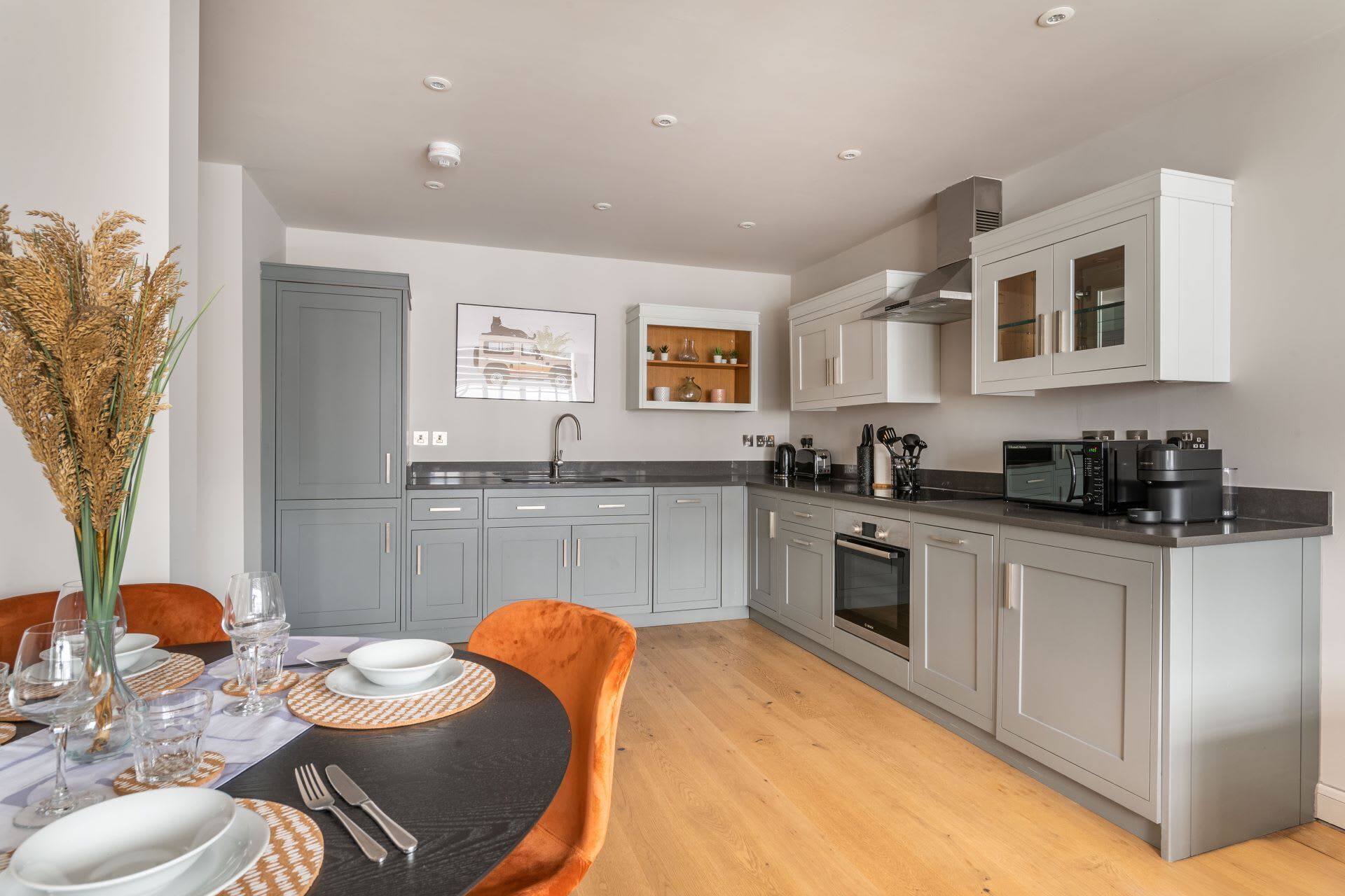 Une cuisine moderne avec des meubles gris, un plan de travail noir et un coin repas comprenant une table ronde et des chaises orange.