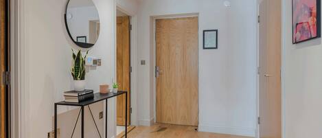 A hallway with a wooden door, a round mirror, a console table with books and a plant, and a framed picture on the wall.