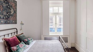 Ein Schlafzimmer mit Bett, Kissen, einem Nachttisch, einer Lampe und einem Fenster mit Blick auf Gebäude.