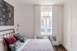 A bedroom with a bed, pillows, a nightstand, a lamp, and a window with a view of buildings outside. - A Starry Starry Night - Borough - by Frankie Says (London)