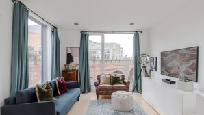 A modern living room with a blue sofa, a brown leather sofa, and a white ottoman. There is a large window with curtains, a clock, and a wall-mounted TV. - The Dream Factory - Borough - by Frankie Says (London)