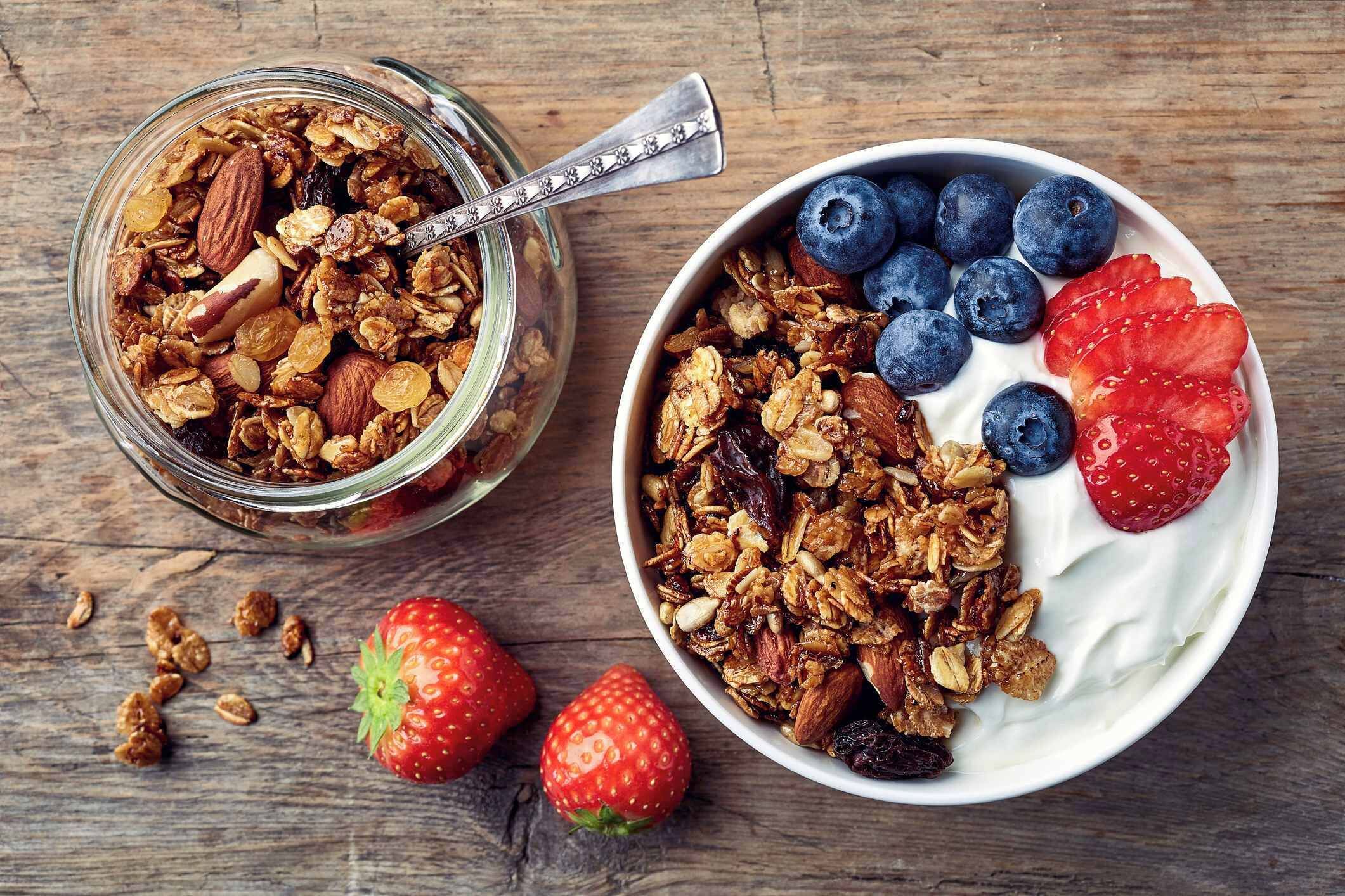 A bowl of granola with blueberries and strawberries, and a jar of granola on a wooden surface.