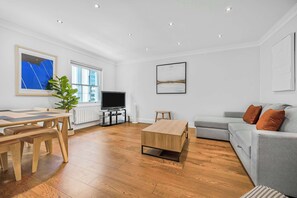 A modern living room with a wooden floor, a grey sofa, a coffee table, a TV, and a dining area with a table and chairs. - A Scholar's Gallery - Bloomsbury - by Frankie Says (London)