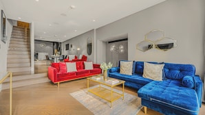 A modern living room with a red and blue sofa set, a coffee table, and a staircase leading to a dining area. - The Royal Mews - Soho - by Frankie (London)