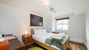 A neatly arranged bedroom with a bed, bedside table, lamp, and a framed cityscape picture on the wall. - Views For Days - Fitzrovia - by Frankie Says (London)