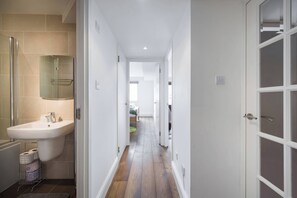 A narrow hallway with a bathroom on the left, a door on the right, and a view into another room at the end. - Views For Days - Fitzrovia - by Frankie Says (London)