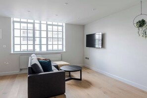 Een moderne woonkamer met een bank, een klein rond tafeltje, een aan de muur bevestigde televisie, en een groot raam dat natuurlijk licht binnenlaat.