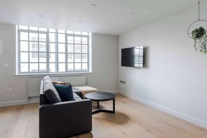 Een moderne woonkamer met een bank, een klein rond tafeltje, een aan de muur bevestigde televisie, en een groot raam dat natuurlijk licht binnenlaat.