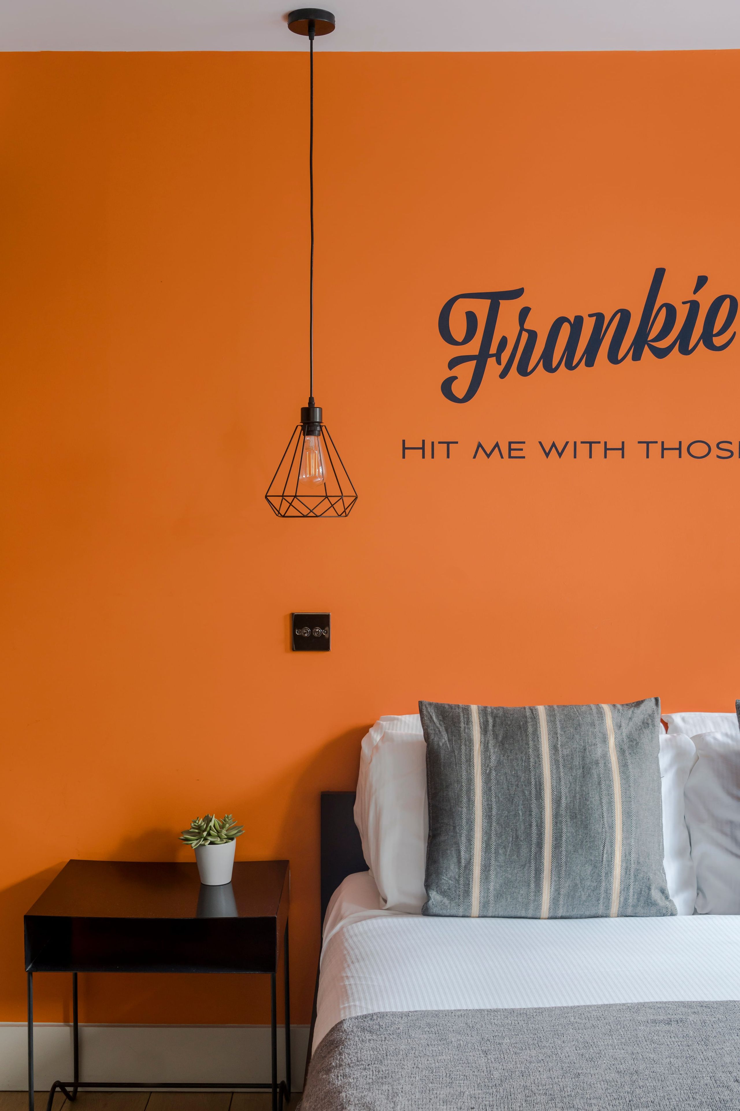 Een kamer met een oranje muur, een bed met kussens, een klein tafeltje met een plant en een hanglamp.
