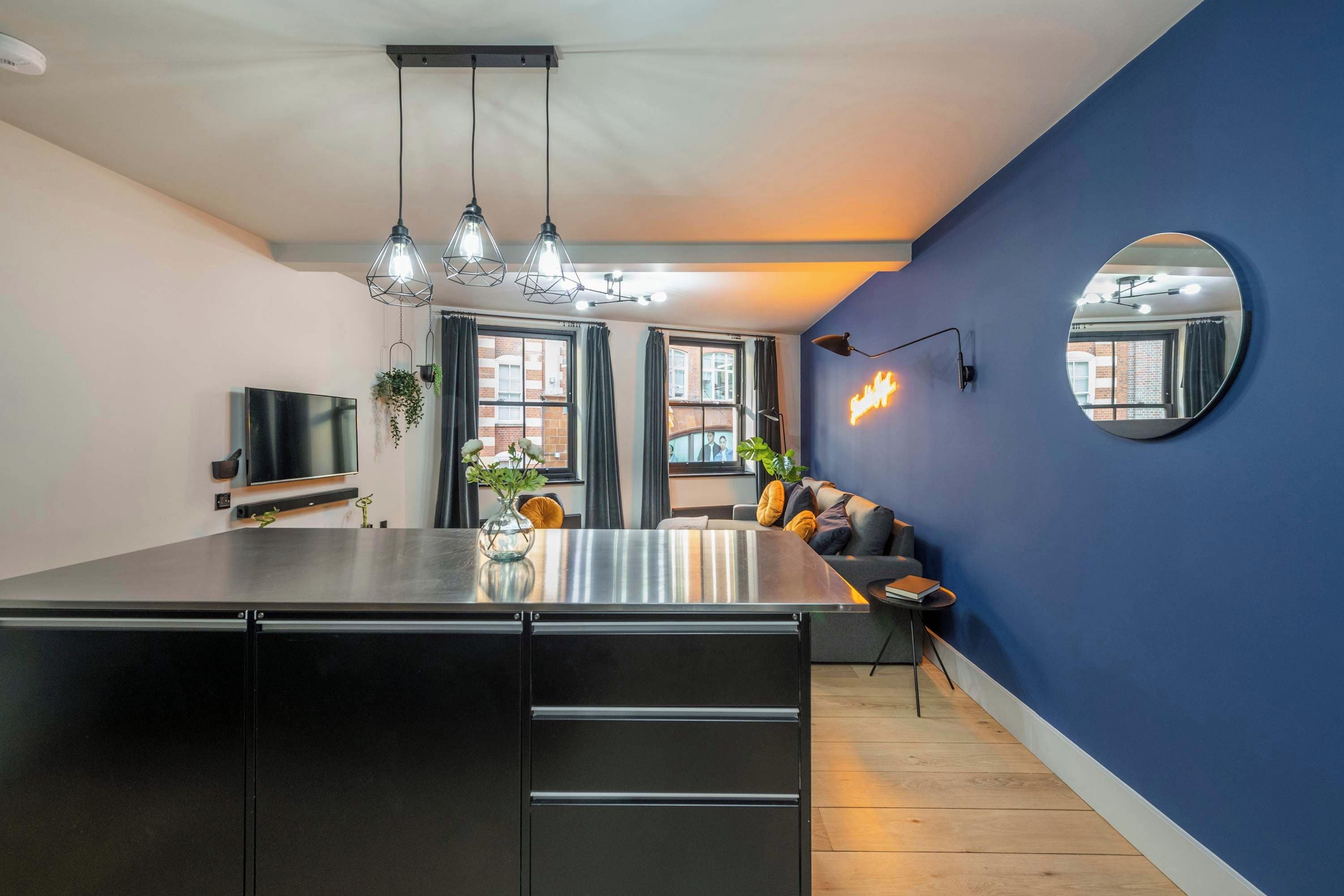 A modern kitchen with a central island, pendant lighting, a flat-screen TV, and a large mirror on the wall.