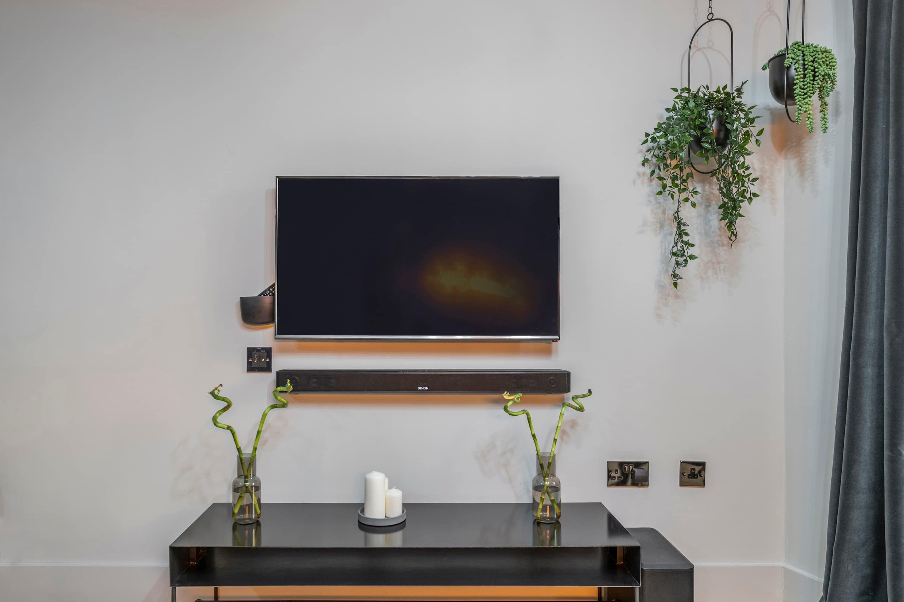 A modern living room with a flat-screen TV mounted on the wall, a black console table with two vases, and hanging plants.