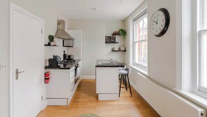 A narrow kitchen with a central island, white cabinetry, a black countertop, and a window with a clock. - The Lipstick Palace - Fitzrovia - by Frankie Says (London)