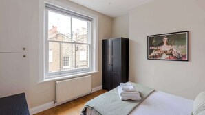 A bedroom with a large window, a radiator, a wardrobe, a bed with a green blanket, and a framed picture on the wall. - The Lipstick Palace - Fitzrovia - by Frankie Says (London)