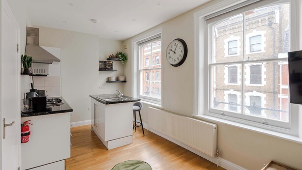 A compact kitchen with a central island, a large window with a view of buildings, a wall clock, and a radiator. - The Lipstick Palace - Fitzrovia - by Frankie Says (London)