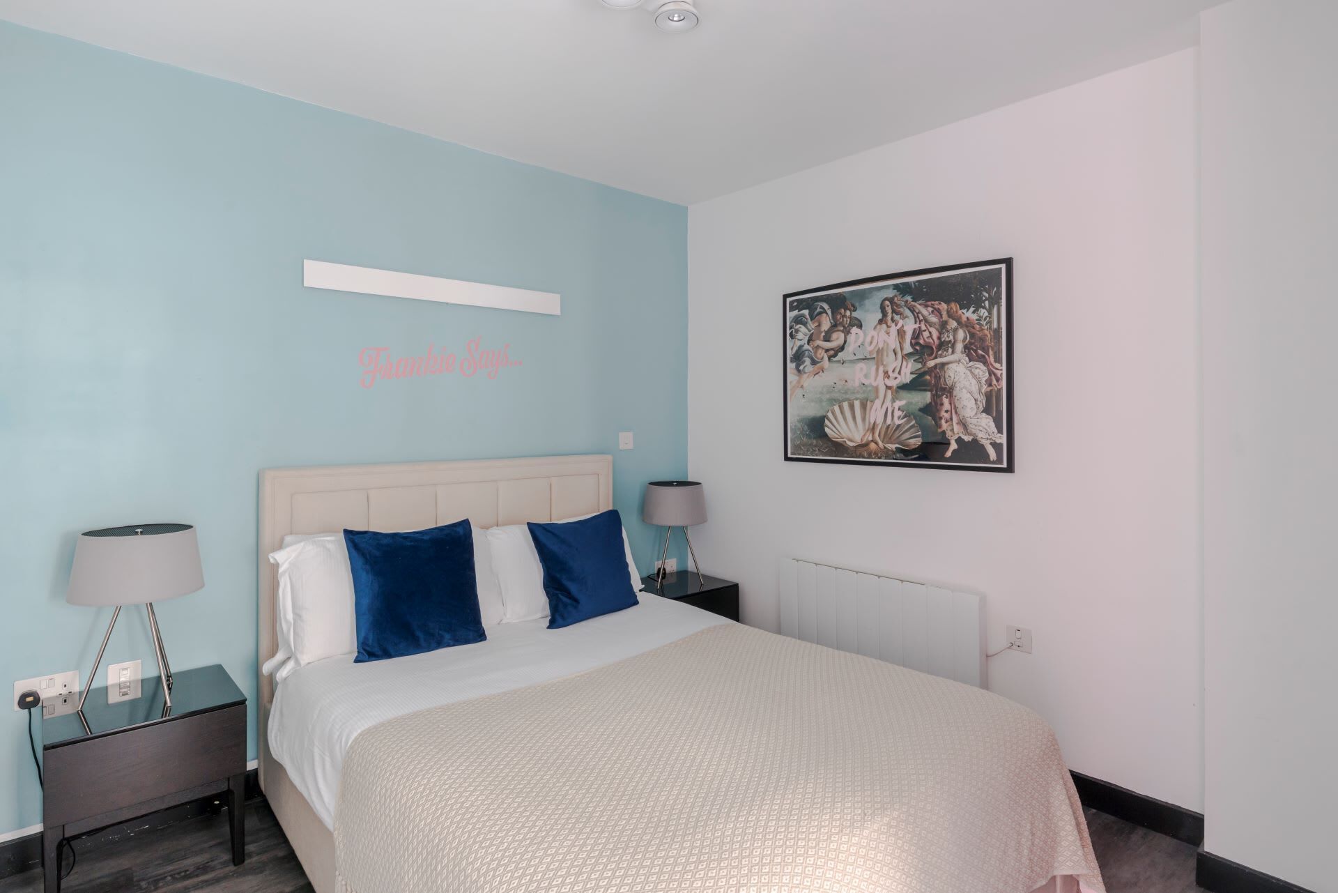 A bedroom with a bed, two bedside tables, a nightstand, a wall-mounted shelf, and a framed picture.