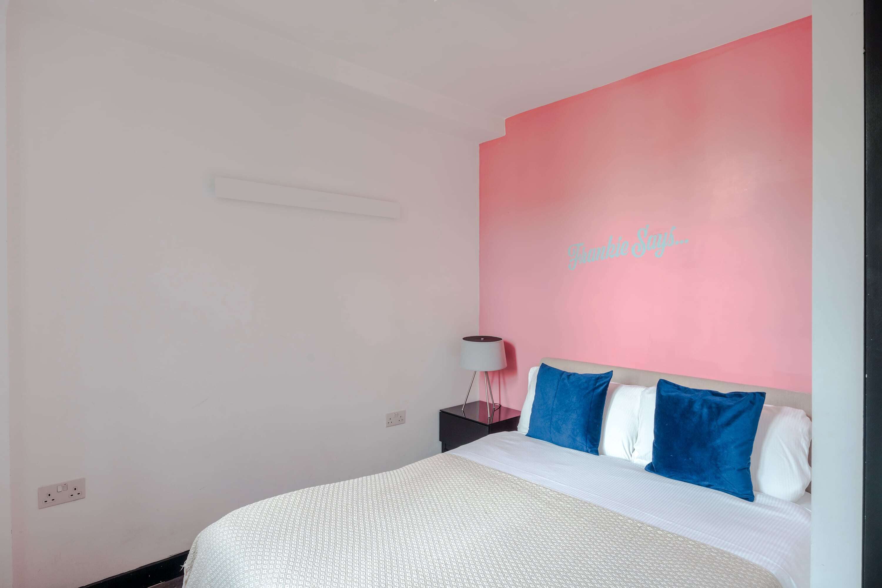 A bedroom with a bed, a nightstand, a lamp, and a pink accent wall.