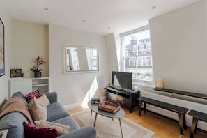 Een woonkamer met een grijze bank, een salontafel, een televisie, een piano en een groot raam met uitzicht op gebouwen.