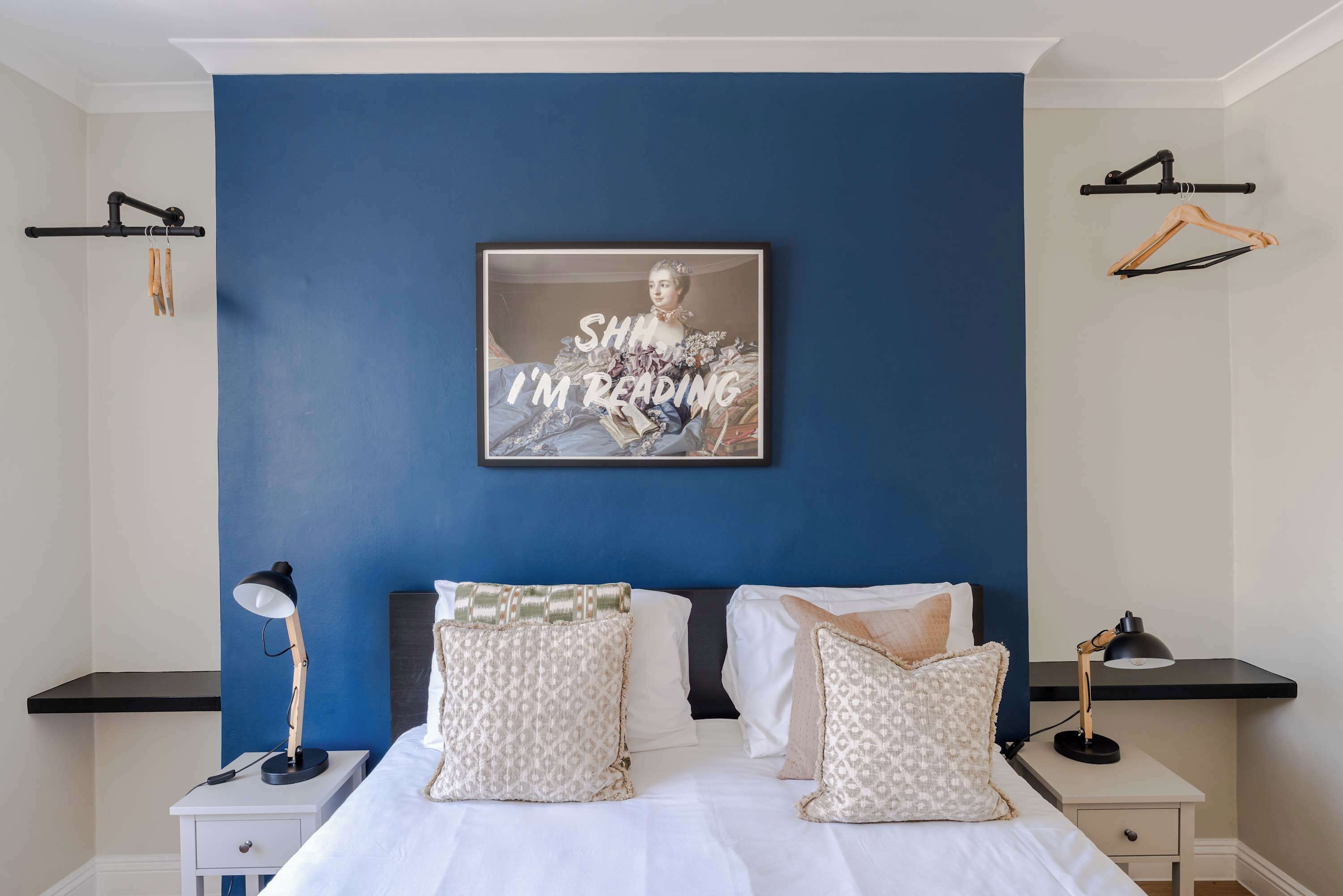Een slaapkamer met een blauwe accentmuur, een lijst met afbeelding, twee nachtkastjes en een bed met gekleurde kussens.