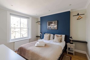 Un dormitorio con un ventanal grande, una cama con ropa de cama blanca, una pared de acento azul y un cuadro enmarcado.
