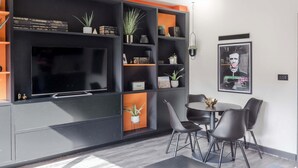Una sala de estar moderna con un televisor de pantalla plana, estanterías negras y naranja, y una zona de comedor con sillas y una mesa.