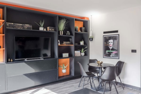Un salon moderne doté d’un téléviseur à écran plat, d’étagères noires et orange, et d’un coin repas avec des chaises et une table.