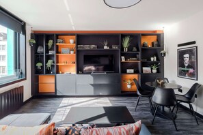 Un salon moderne avec un téléviseur, des étagères noires et orange, un coin repas avec des chaises et une fenêtre.