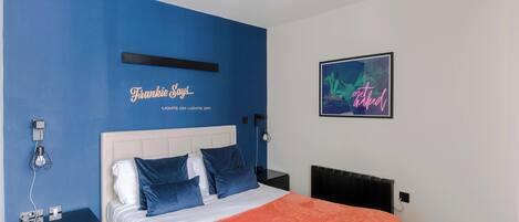Un dormitorio con cama, mesitas de noche, una lámpara fijada a la pared y un cuadro enmarcado.
